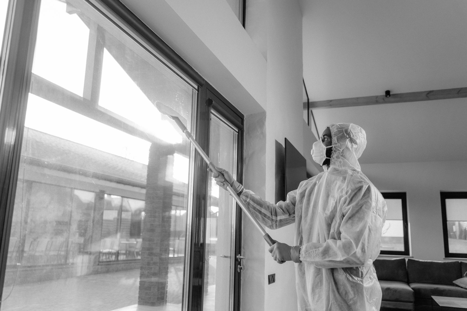 A man in a protective suit is spraying a window in a living room.