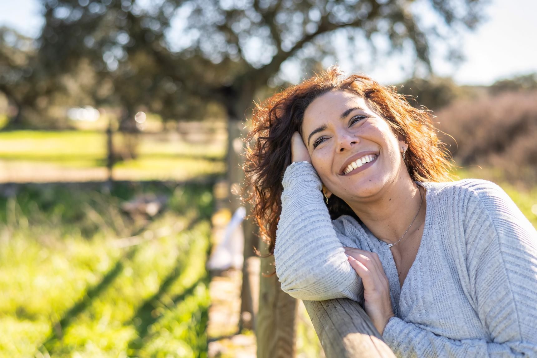 A woman is leaning on a wooden fence in a park and smiling.
