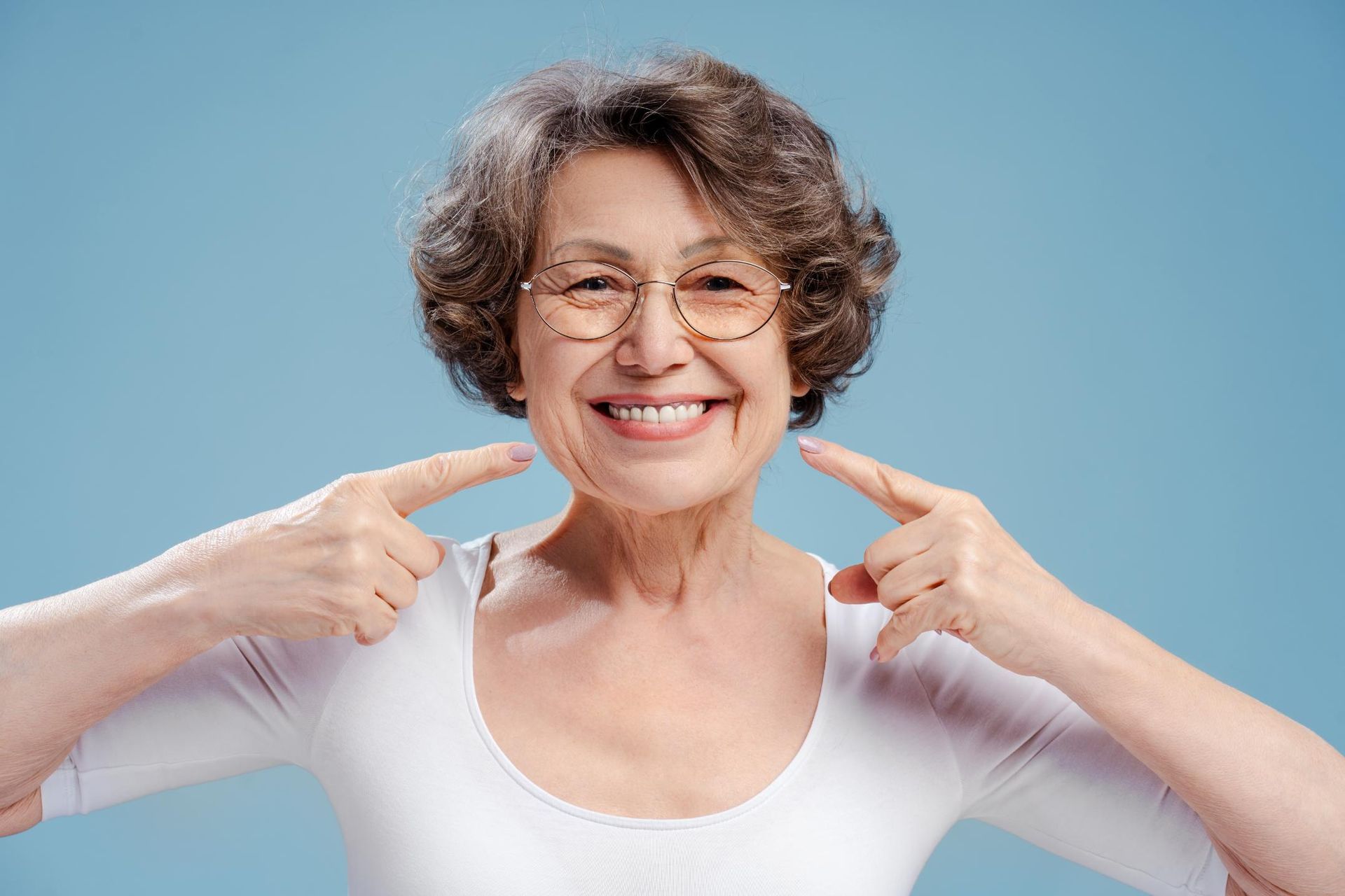 An elderly woman is smiling and pointing at her teeth.