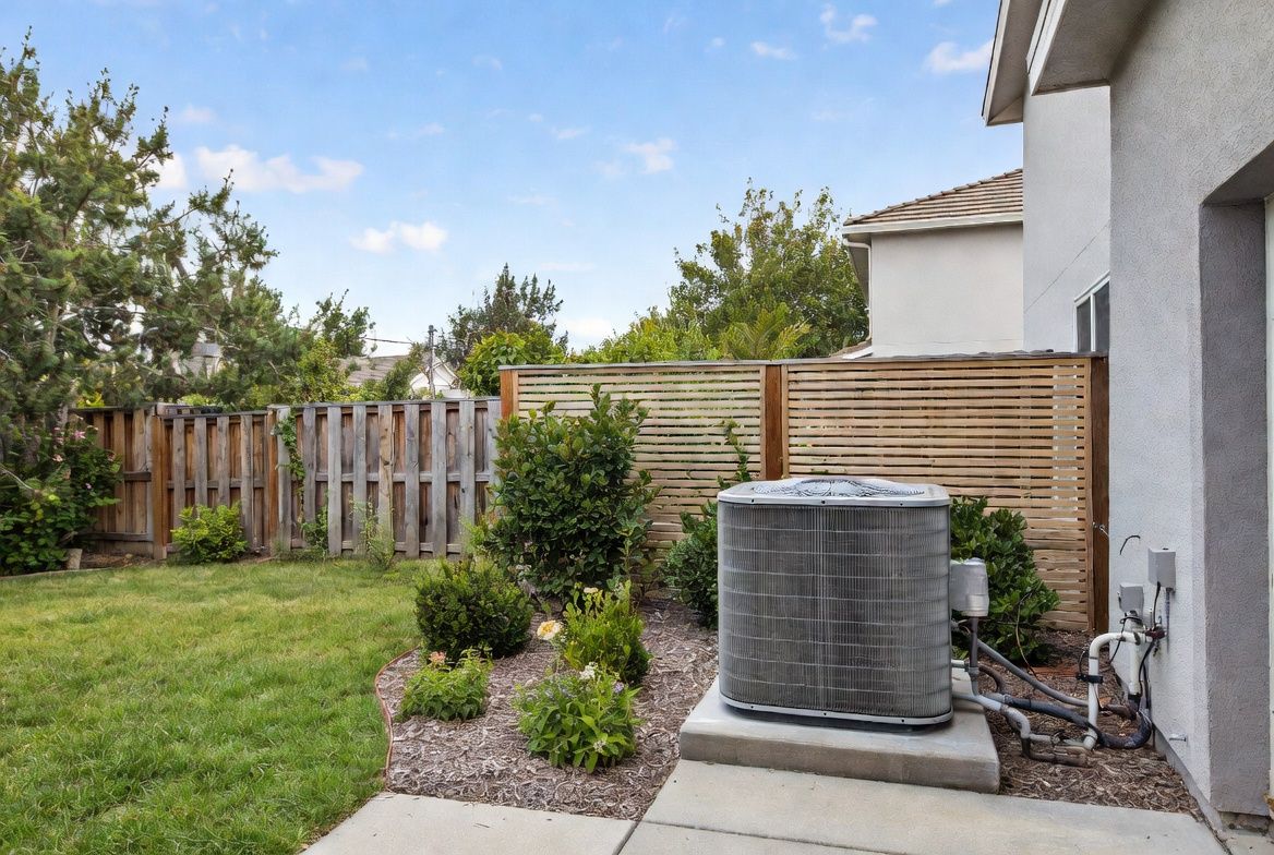 Backyard patio with an outdoor AC unit, lawn, shrubs, wooden fence, and house exterior