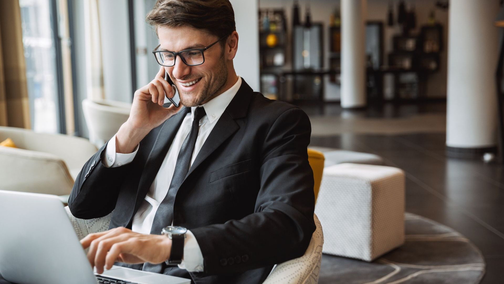 A man in a suit is sitting in front of a laptop computer while talking on a cell phone.