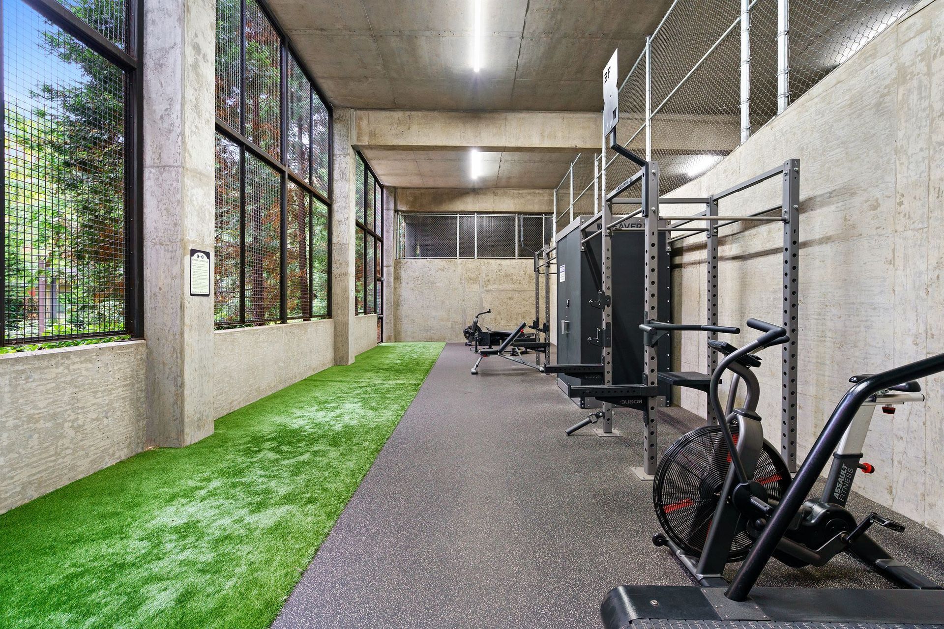 Industrial-style community gym with weight racks, cardio machine, and green turf by windows.