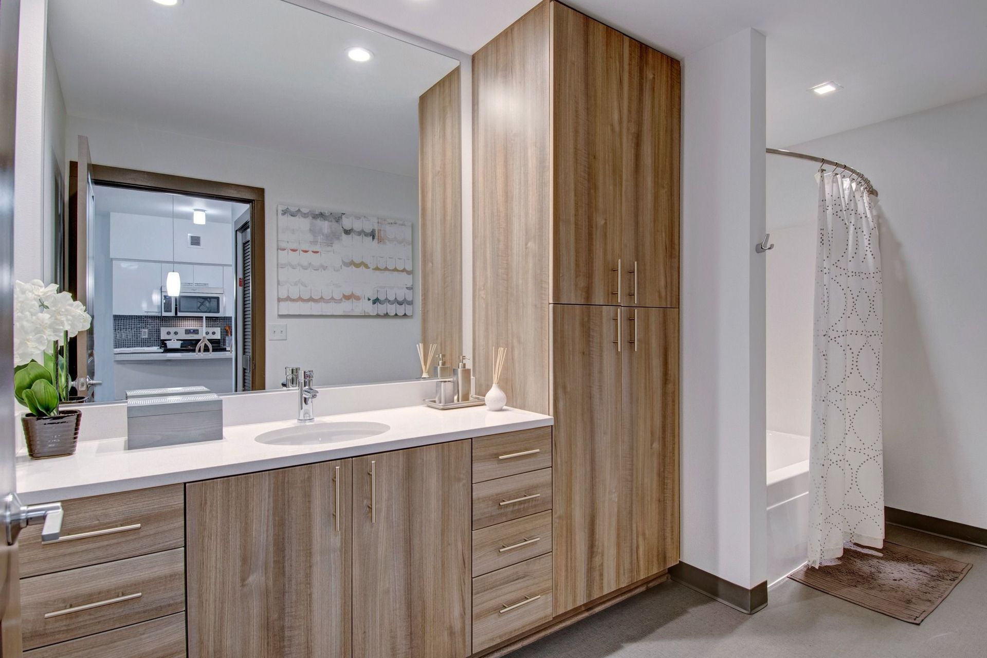 Modern apartment bathroom with wood grain vanity, white countertop, large mirror, and a shower curtain at Tessera at Orenco Station.