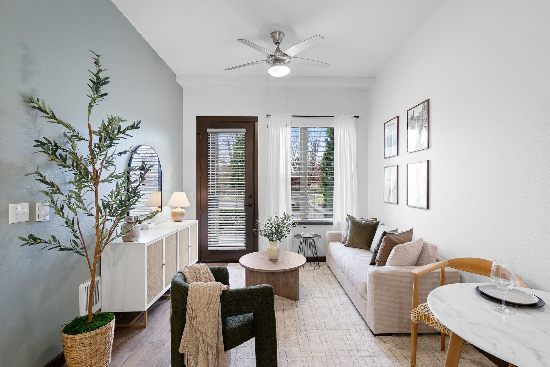 Bright living room with beige sofa, round coffee table, ceiling fan, and glass door with blinds at Tessera at Orenco Station.