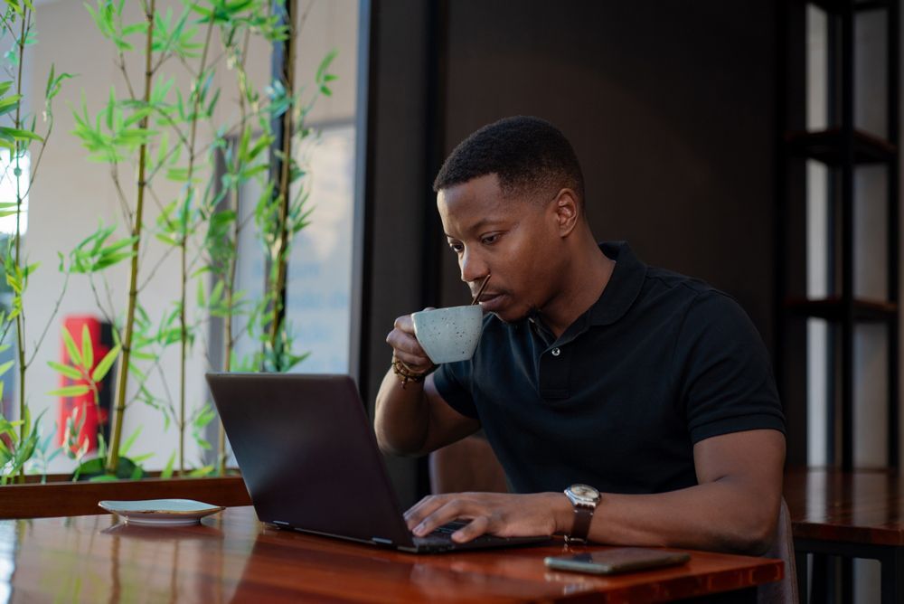 A man is sitting at a table using a laptop computer while drinking a cup of coffee.