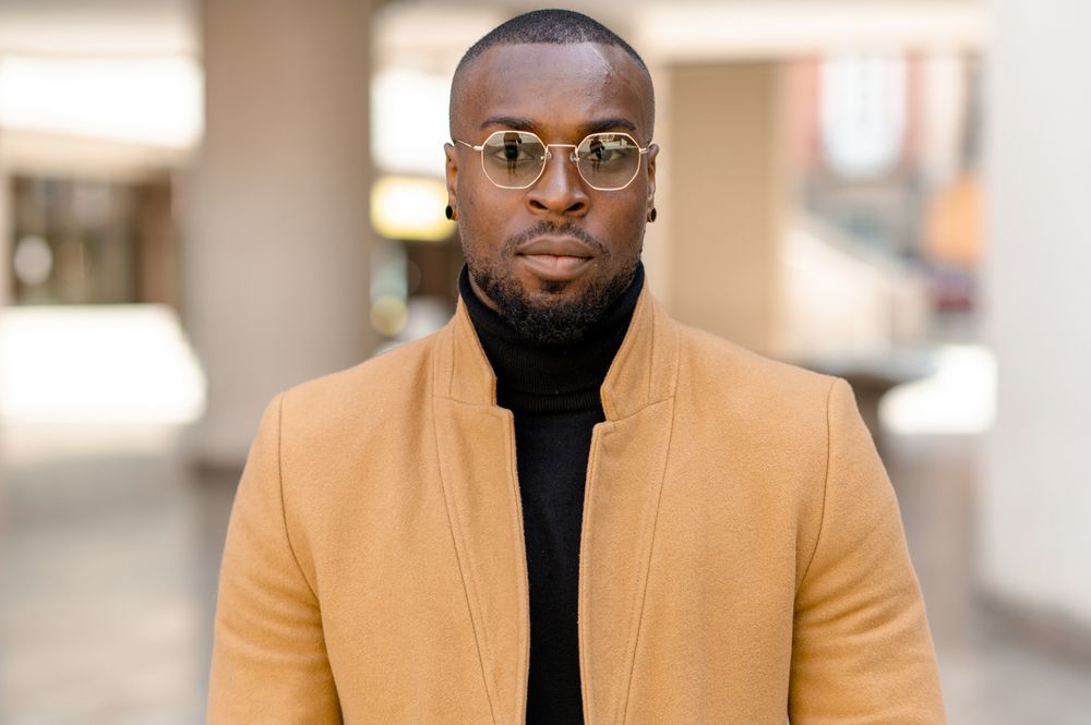 A man wearing glasses and a tan coat is standing in a hallway.