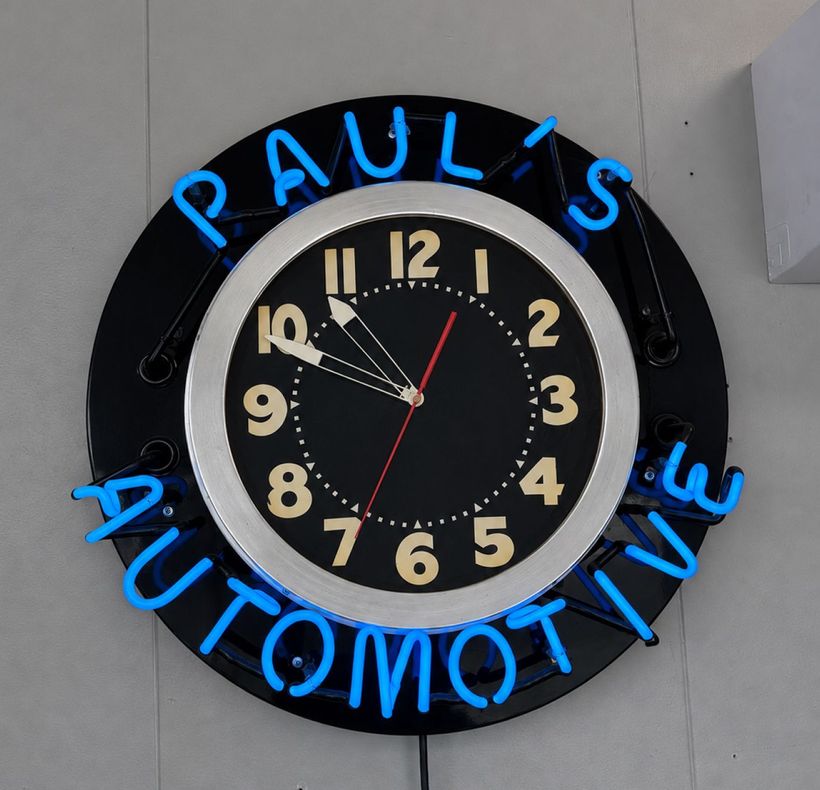 Round black clock sign with blue neon-style text reading “PAUL’S AUTOMOTIVE” on a wall | Paul's Automotive