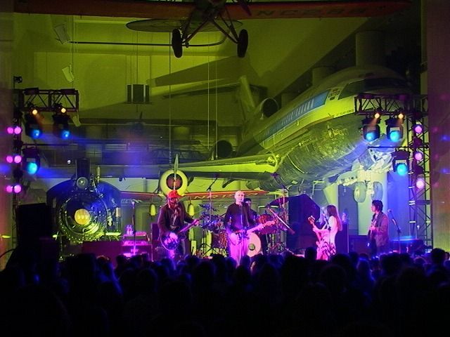 Live band performing onstage under purple and green lights, with a crowd silhouetted in front.