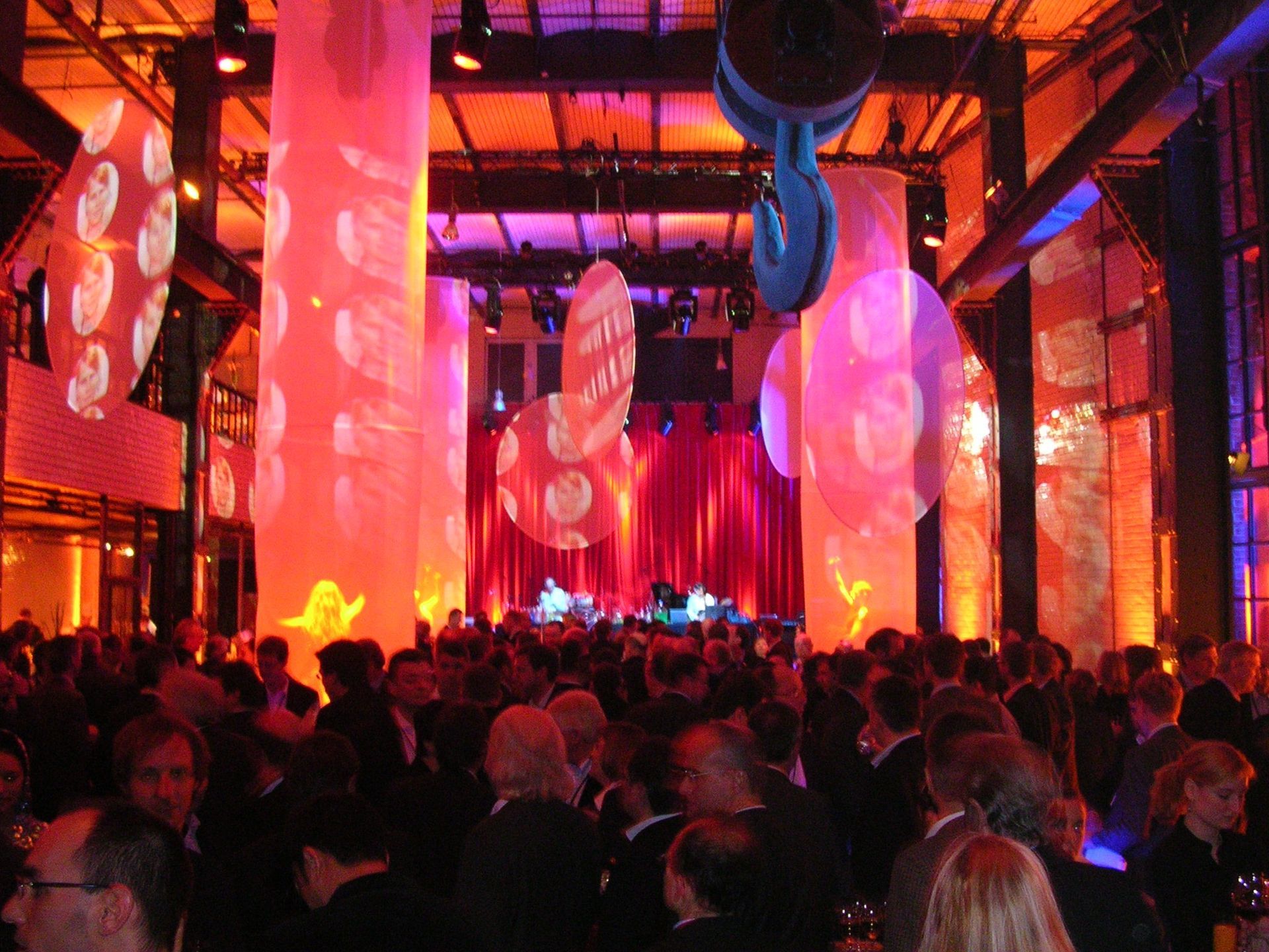 Crowded indoor event with colorful red and pink lighting, hanging fabric columns, and a stage in the distance