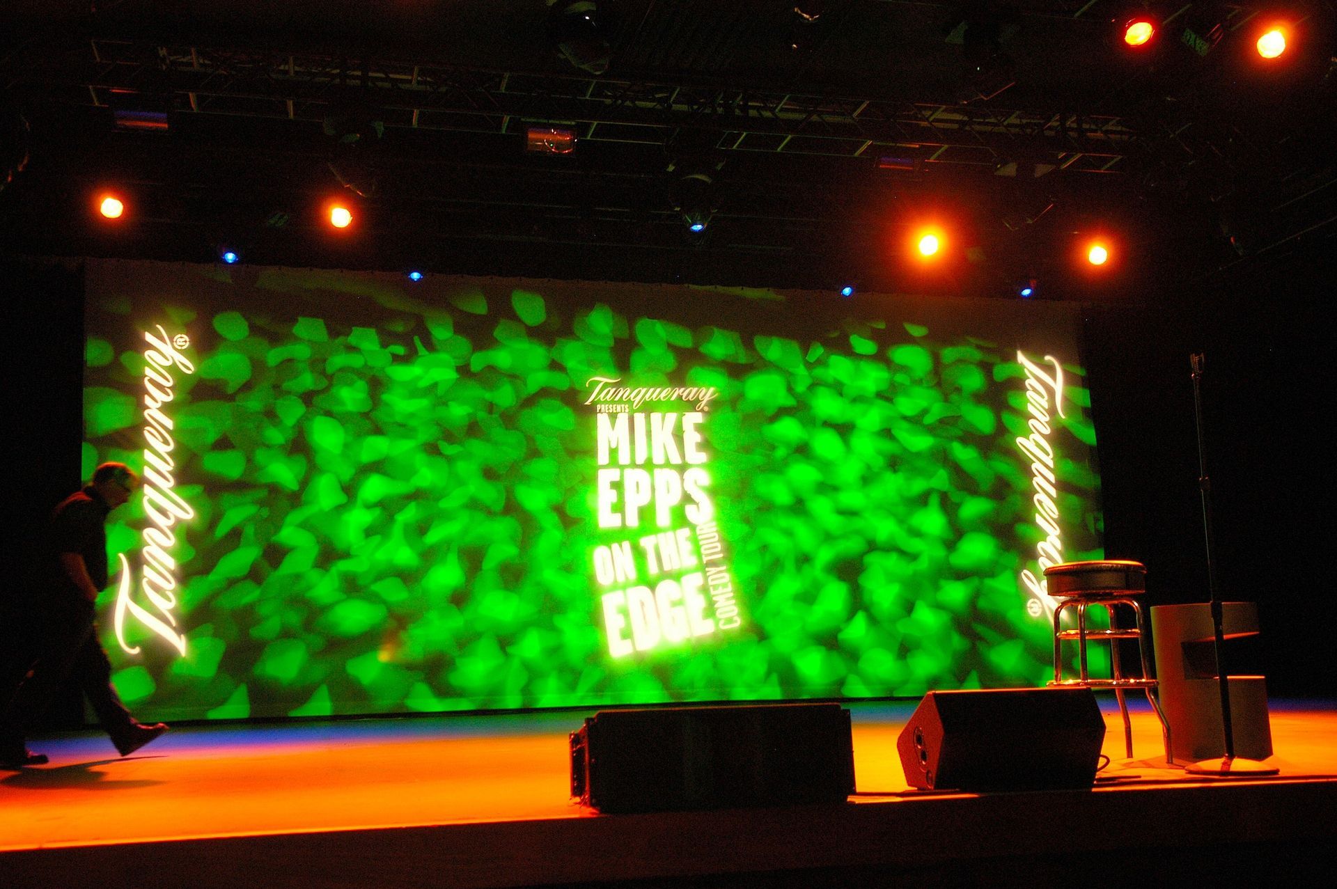 Green-lit stage with large screen reading “MIKE EPPS ON THE EDGE” and a chair at right.