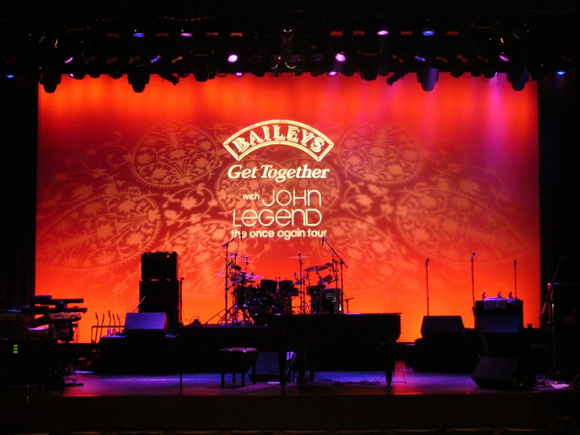 Concert stage with red-lit backdrop, drum kit, microphones, and musical equipment before a live event