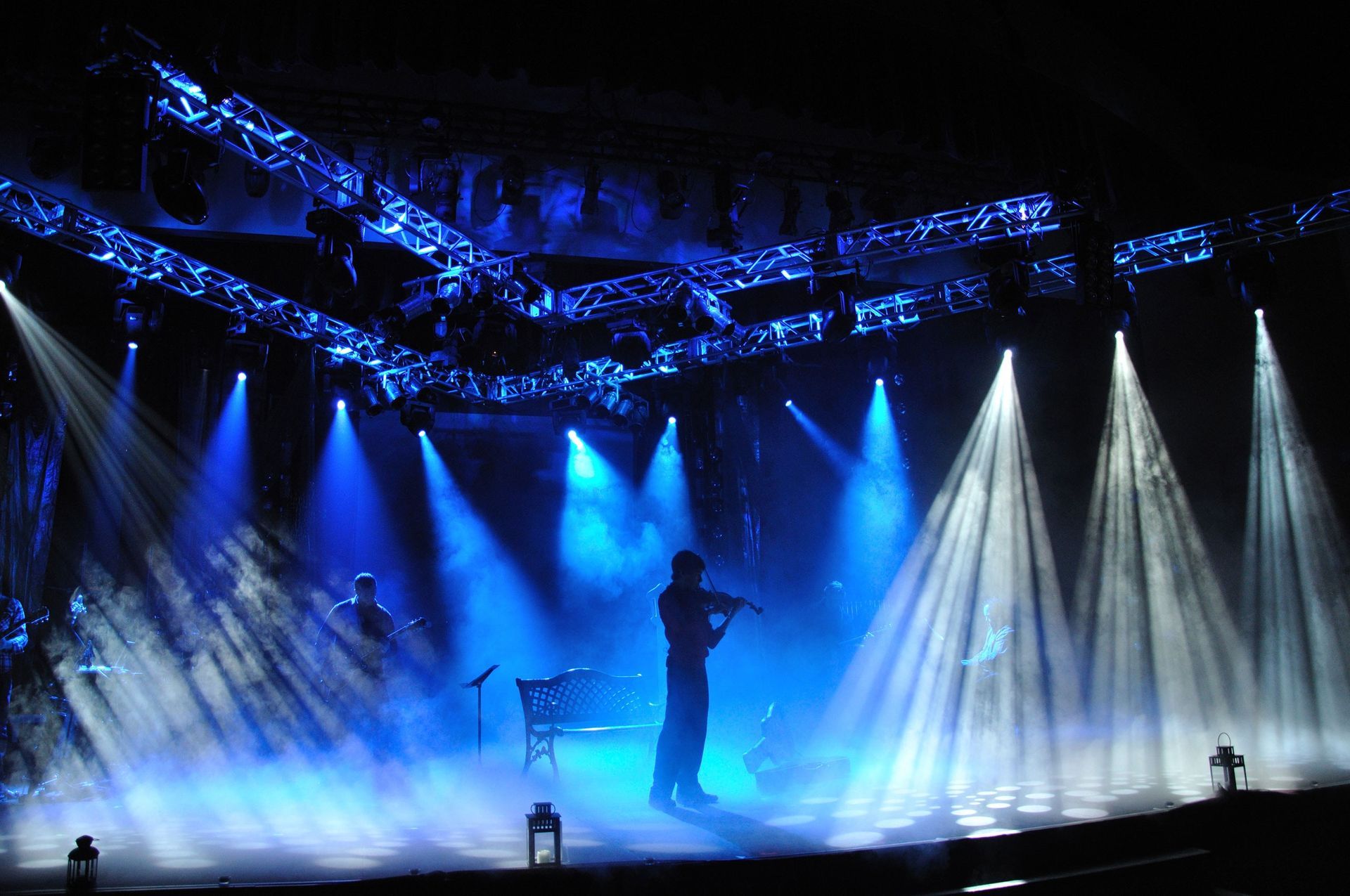 Blue-lit concert stage with a silhouetted performer and bright white spotlights through haze