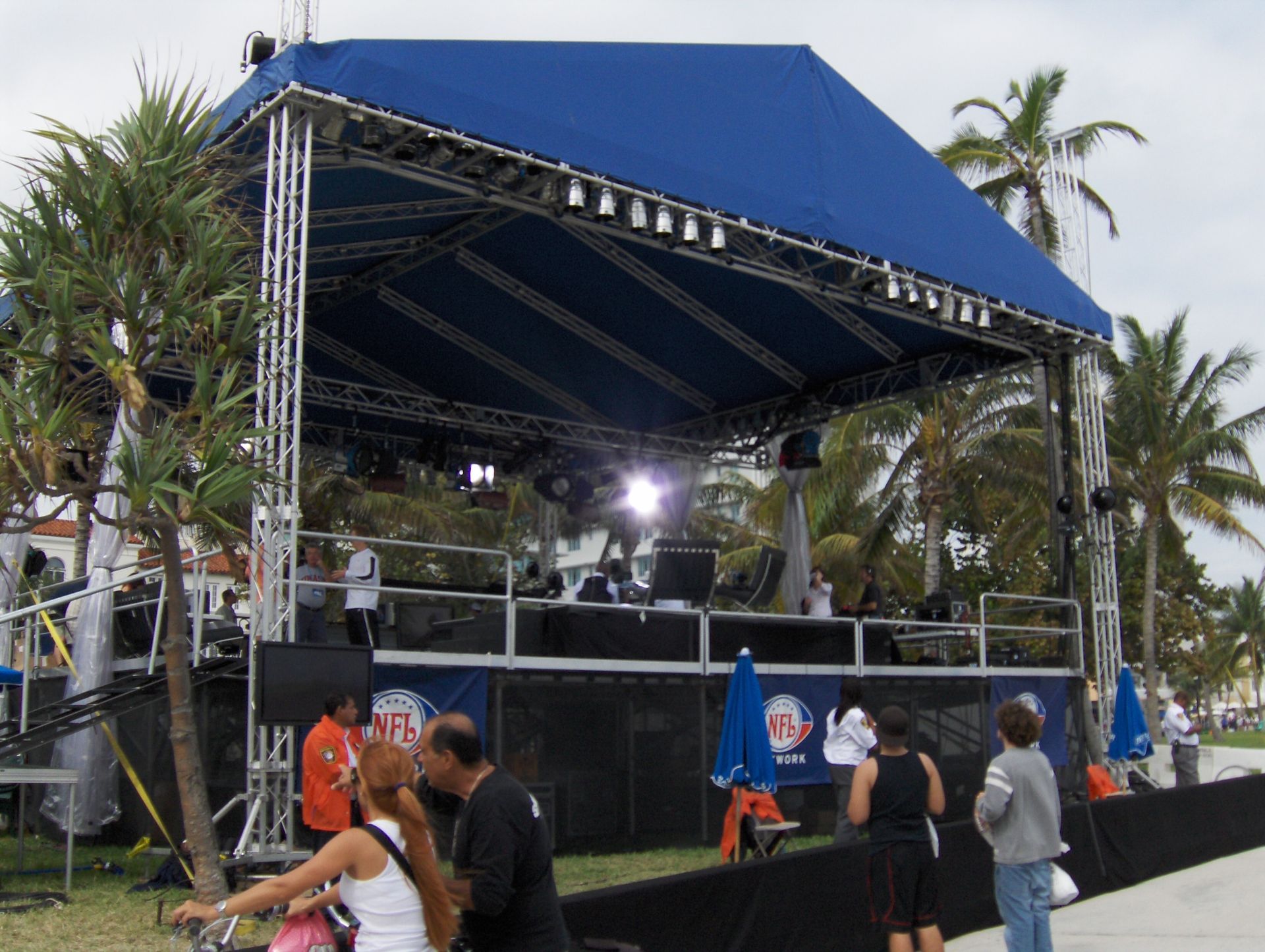 Outdoor stage with blue canopy, lighting rig, and a few people standing nearby under palm trees.