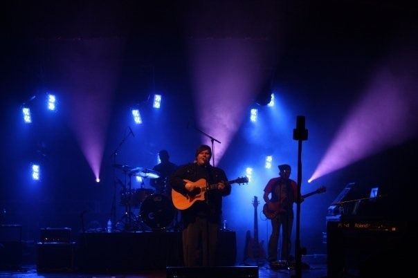 Band performing on a dark stage with blue and purple lights, guitarist centered and drummer in back.