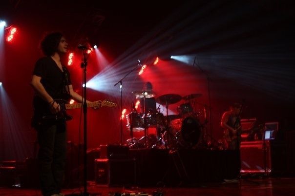 Band performing on a red-lit stage, with guitarist at left and drummer center amid bright spotlights.