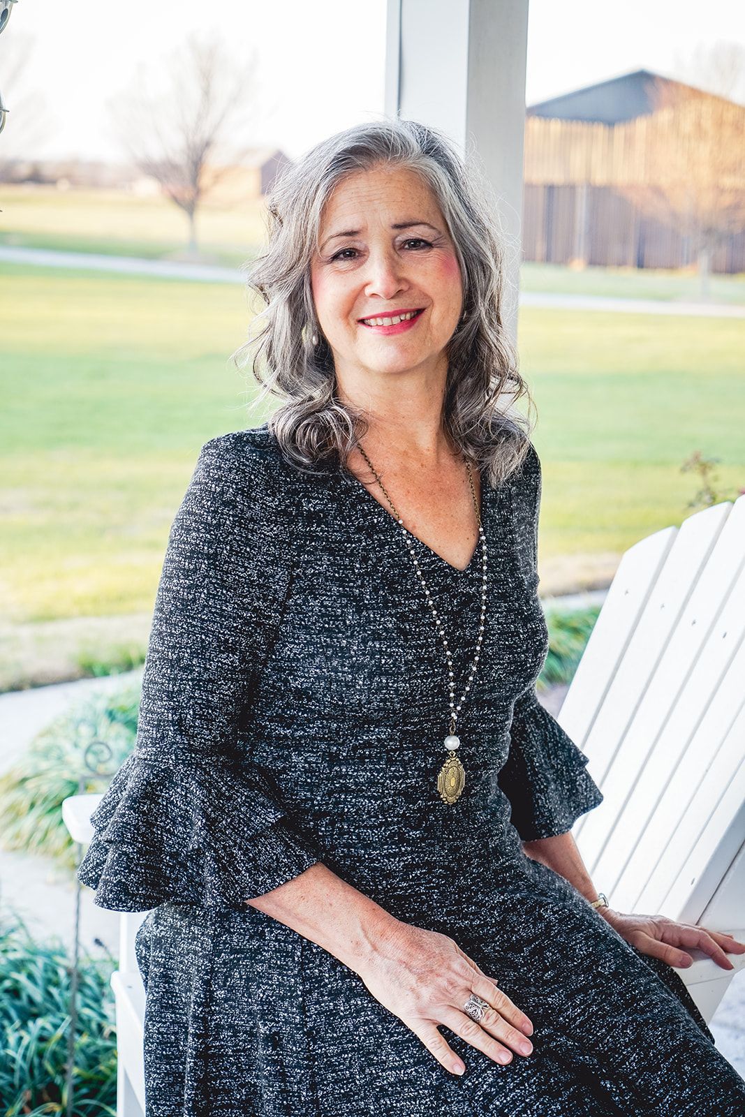 Author Sandra Wiseman Smith in dark gray dress with long pendant sitting on white Adirondack  chair.