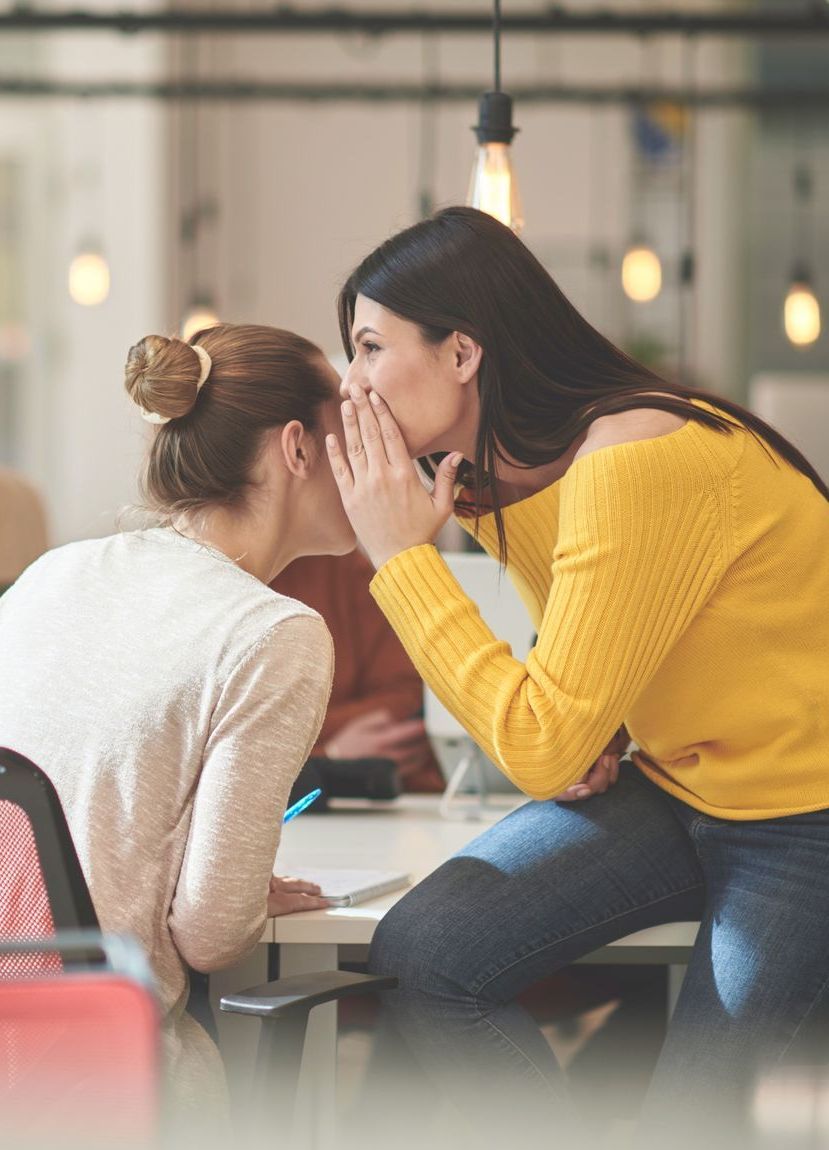 Dark-haired young woman in mustard yellow top whispering w/ her hand cupped to another young woman in white top facing away. 