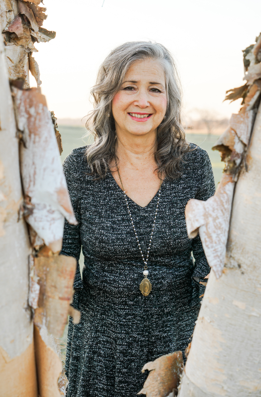 Author Sandra Wiseman Smith smiling and wearing dark gray tweed dress and long pendant standing between two birch trees.
