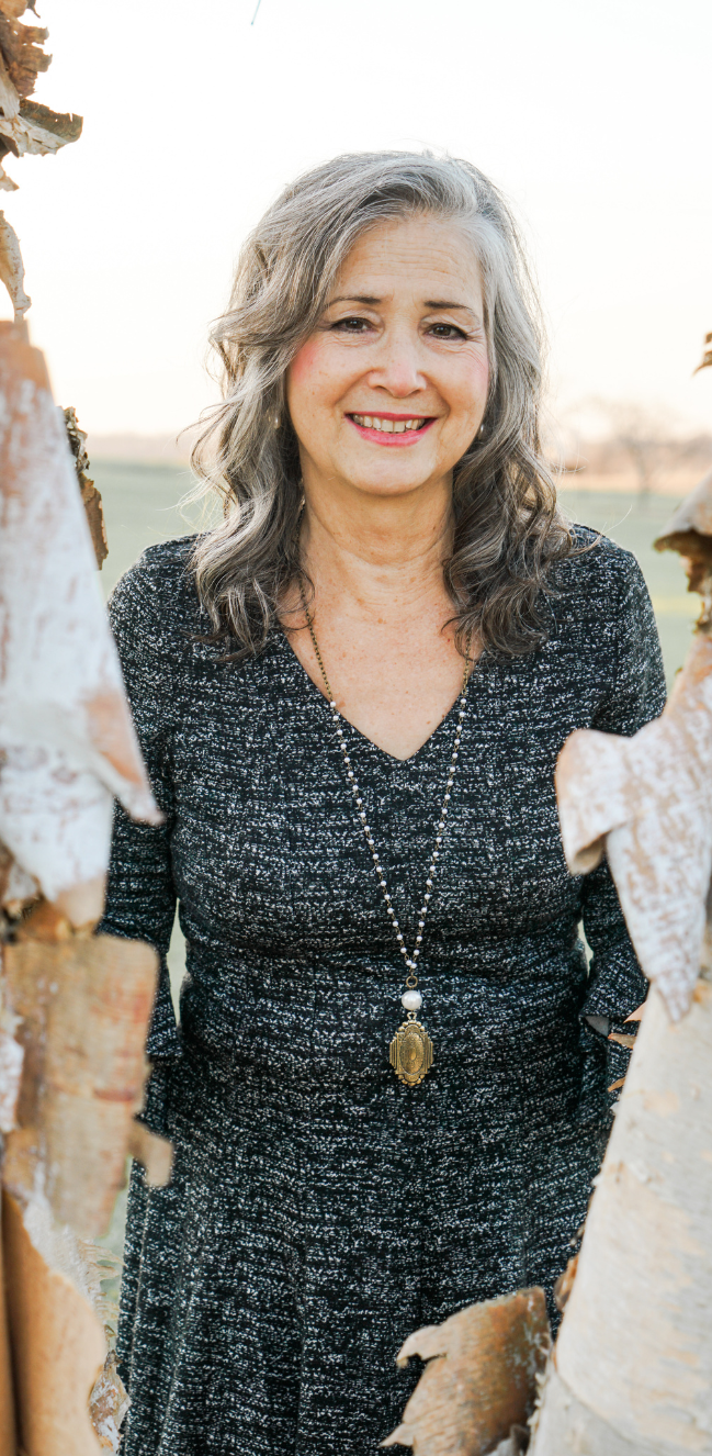 Author Sandra Wiseman Smith smiling and wearing dark gray tweed dress and long pendant standing between two birch trees.