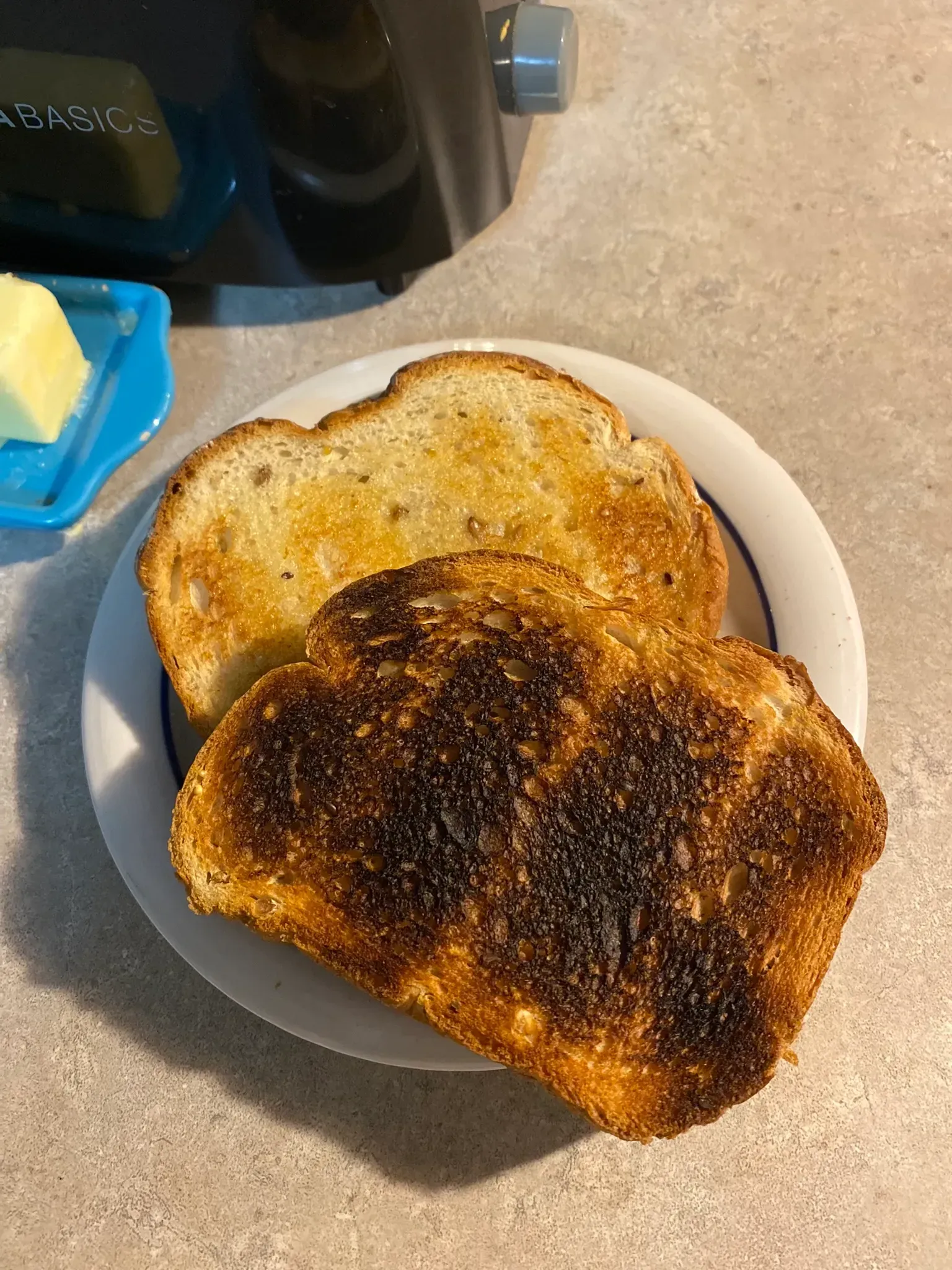Two pieces thick-sliced toast--one burnt, the other buttered and golden, on beige ceramic plate next to teal butter dish.