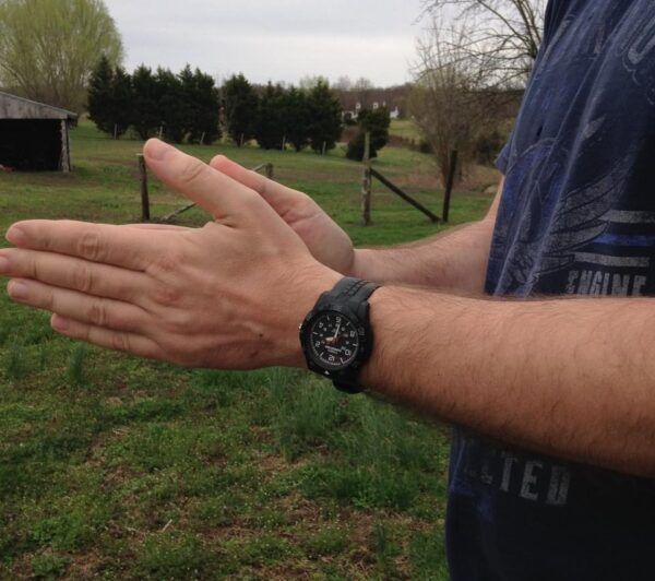 Young man with large black watch and navy blue shirt rubbing his hands together out doors with fence and shed in background.