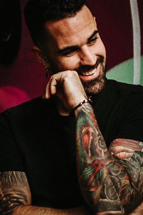 Persona sonriente con una camiseta negra y los brazos tatuados, apoyando la barbilla en la mano ante un fondo colorido desenfocado