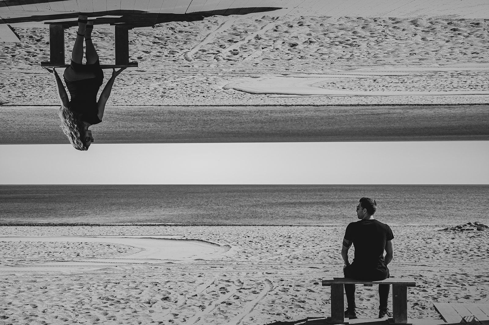 Una persona sentada en un banco frente a una playa tranquila, con la figura invertida reflejada encima en una escena en blanco y negro como en un espejo