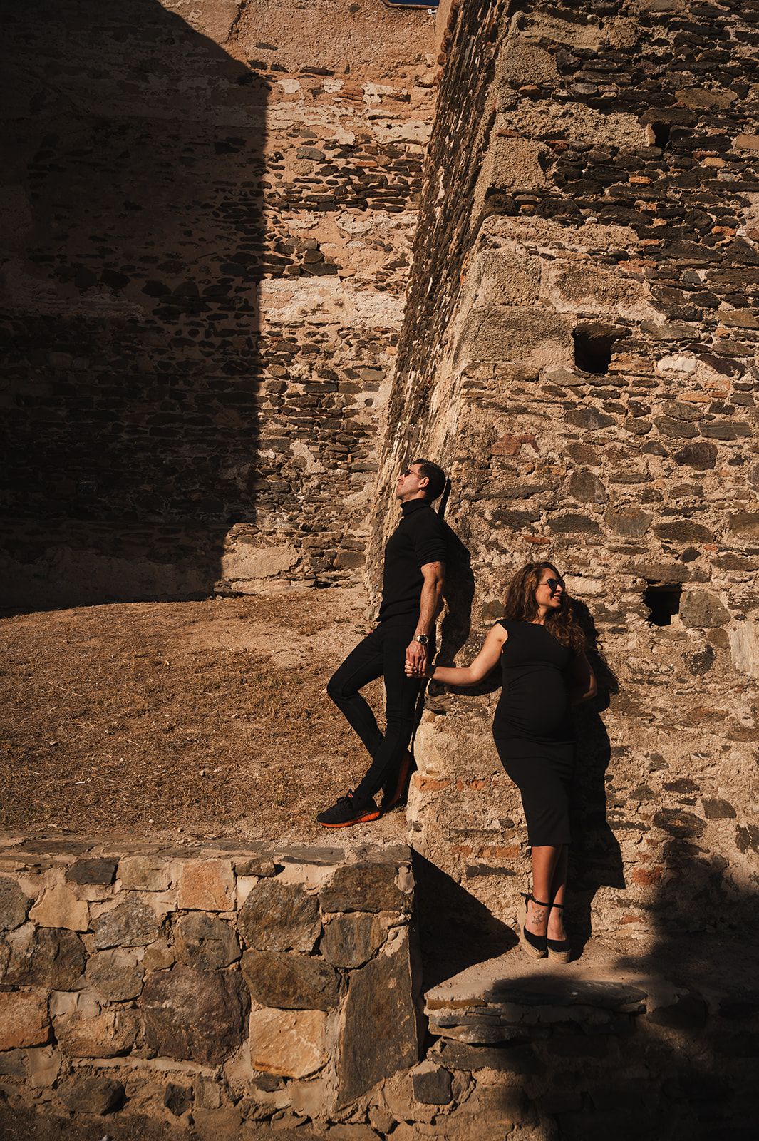 Dos personas de pie junto a un muro de piedra iluminado por el sol, en el patio de un castillo, tomadas de la mano.
