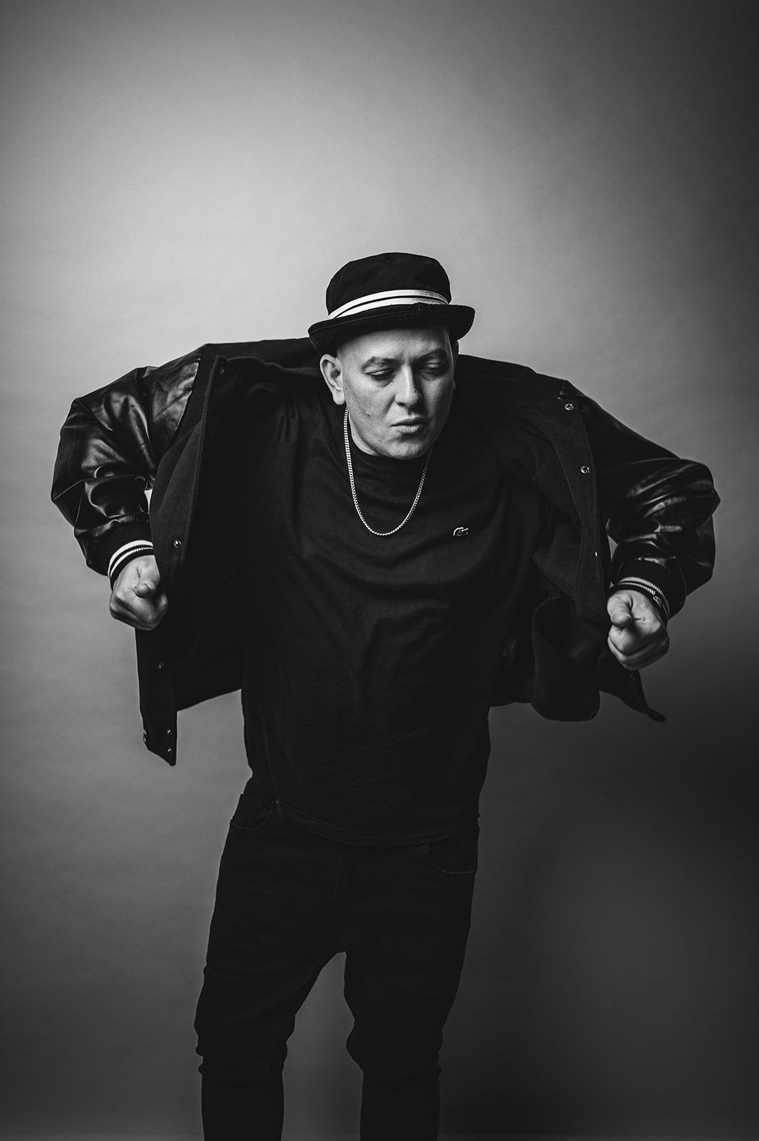 Retrato en blanco y negro de una persona con sombrero y chaqueta, inclinada hacia delante sobre un fondo liso.