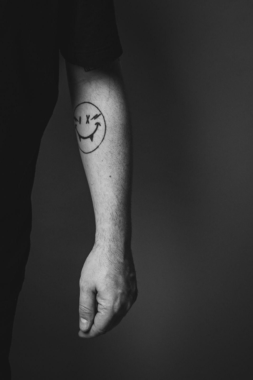 Un antebrazo tatuado con una cara sonriente en la piel, colgando sobre un fondo oscuro.