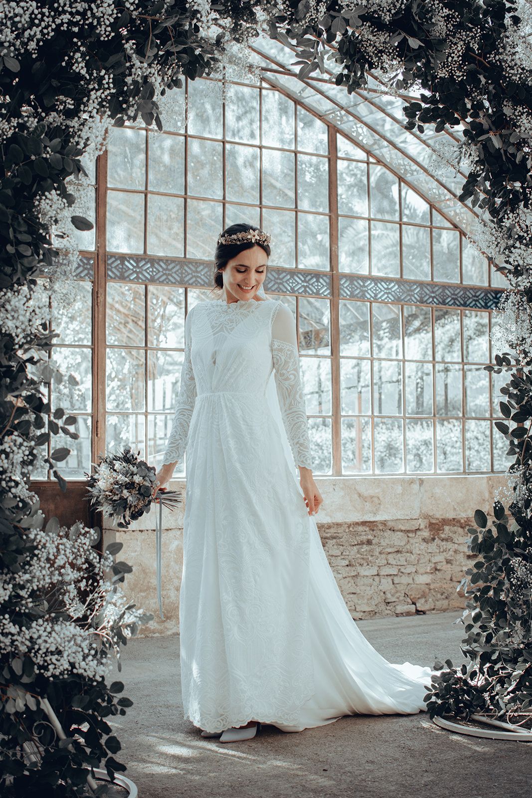 Mujer con un vestido blanco junto a una ventana de cristal arqueada, en un entorno rústico y floral