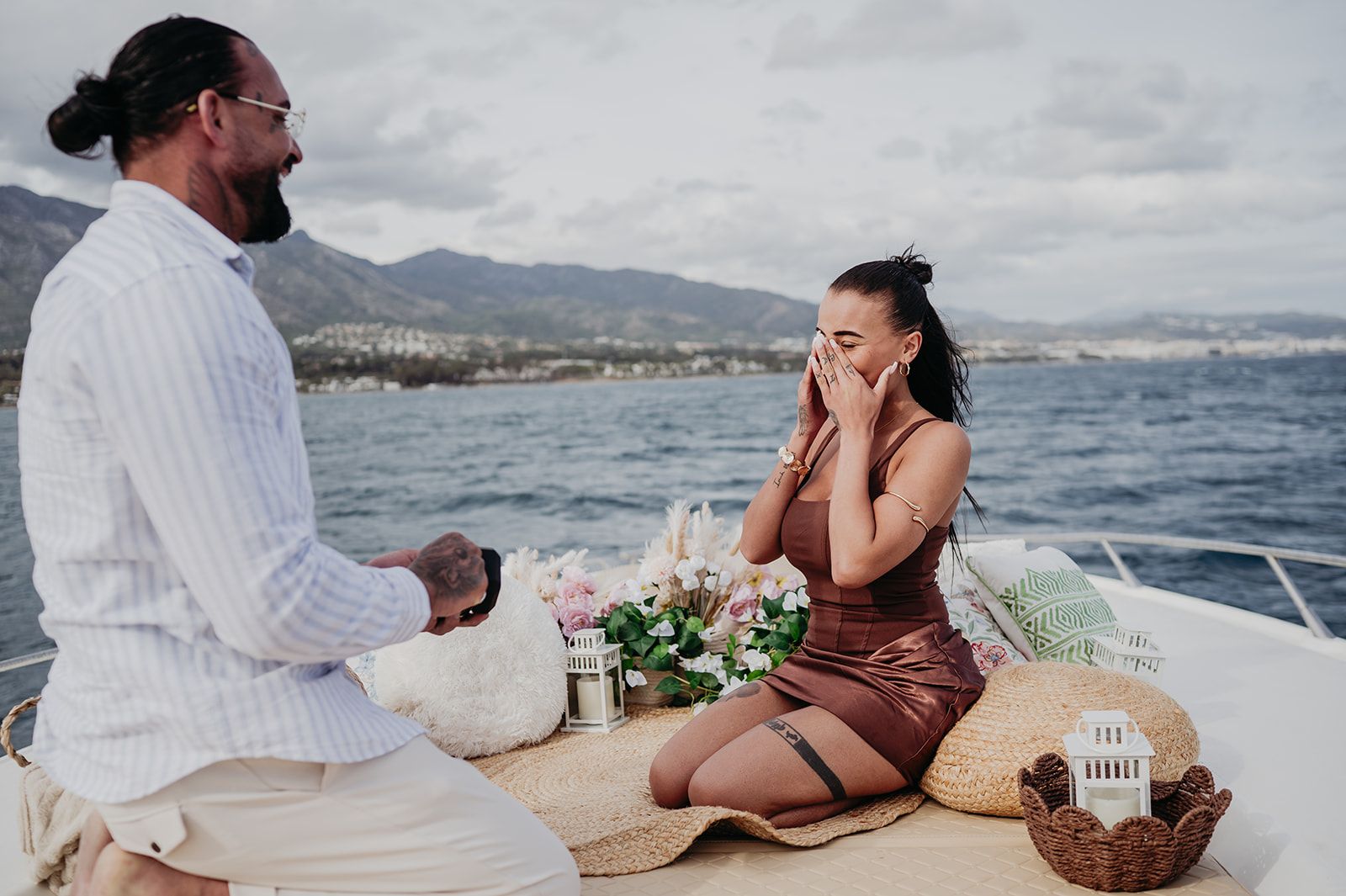 Una pareja de picnic en un barco, una persona arrodillada y la otra sentada sobre una manta con flores, junto a aguas tranquilas y montañas