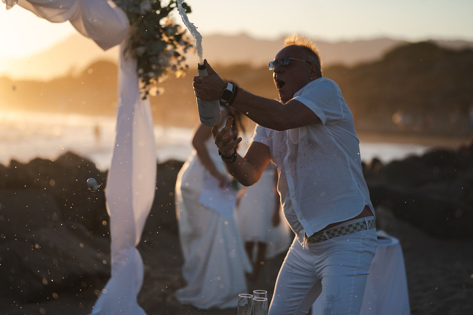 Gente celebrando al atardecer, bailando bajo un arco de cintas blancas decorado con flores.