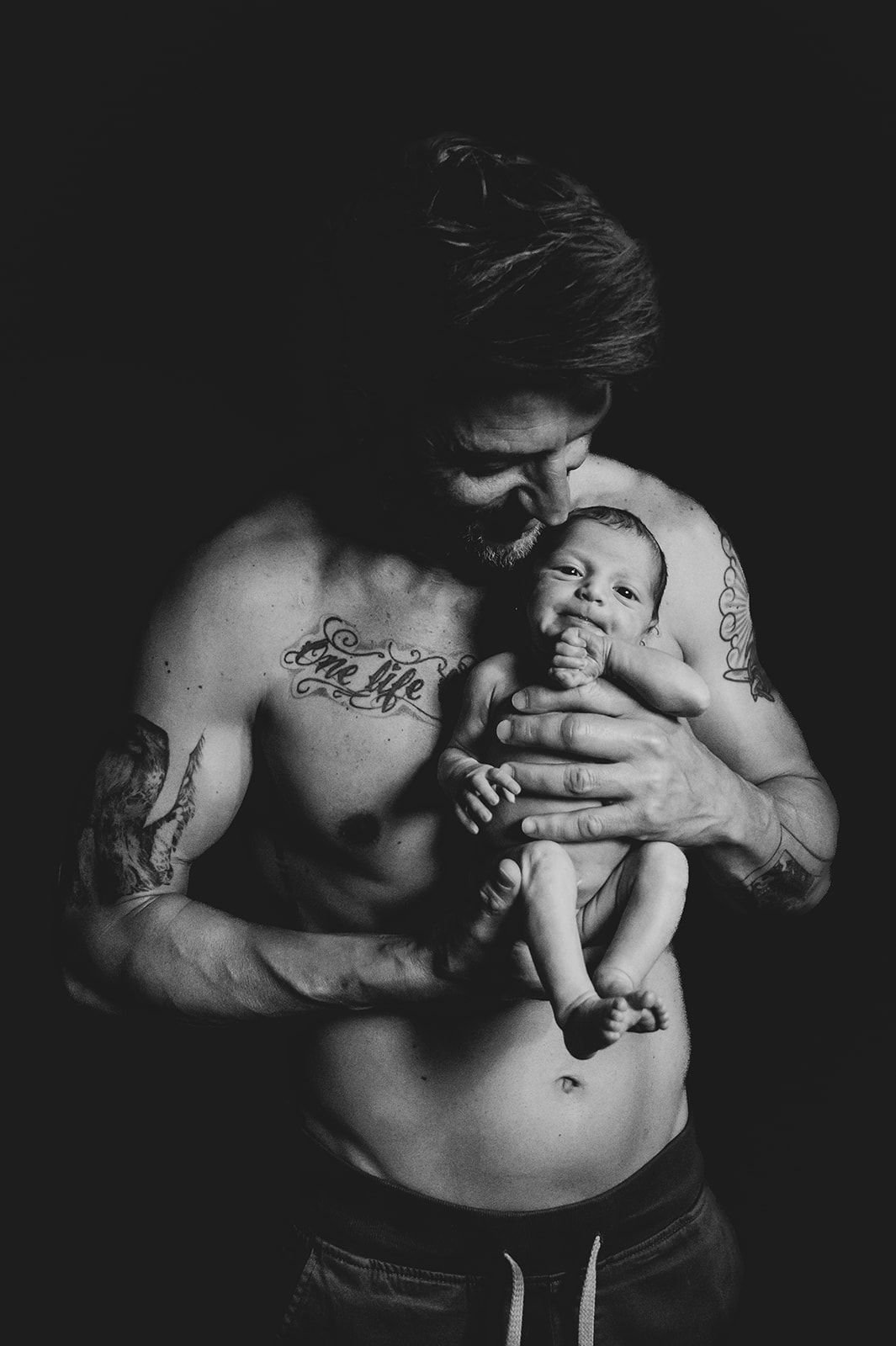 Una persona sin camiseta sostiene a un bebé sonriente sobre un fondo oscuro, en blanco y negro.