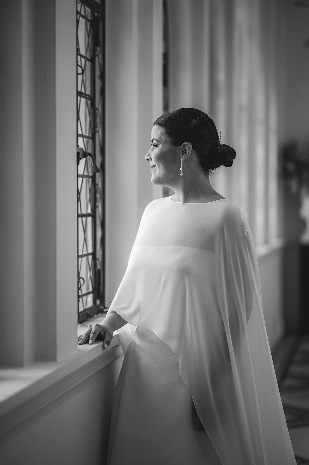 Una mujer con un vestido blanco vaporoso contempla el exterior por una ventana en una escena en blanco y negro con luz tenue