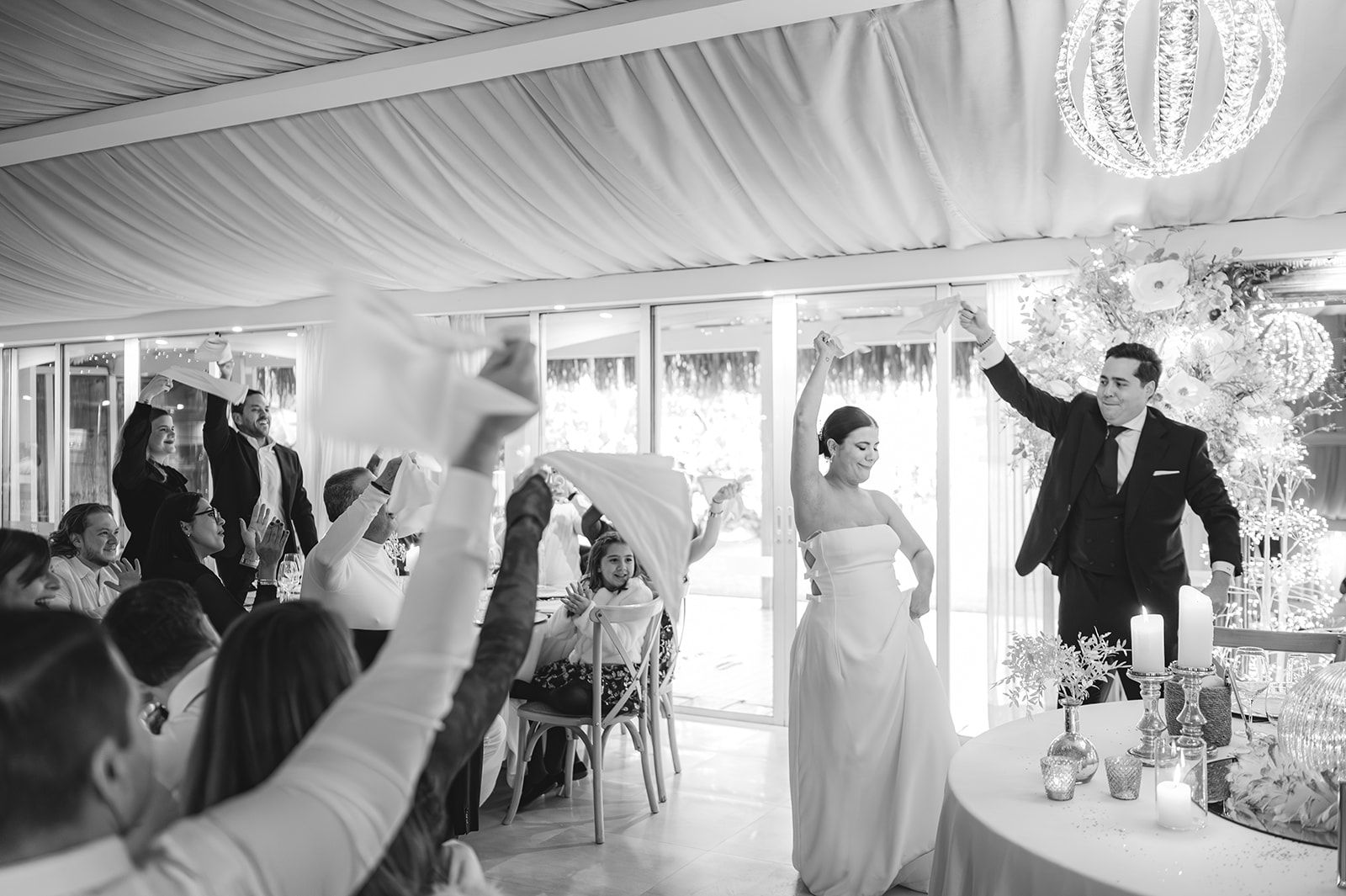 Recepción de boda con los novios brindando bajo luces colgantes en el techo y los invitados vitoreando