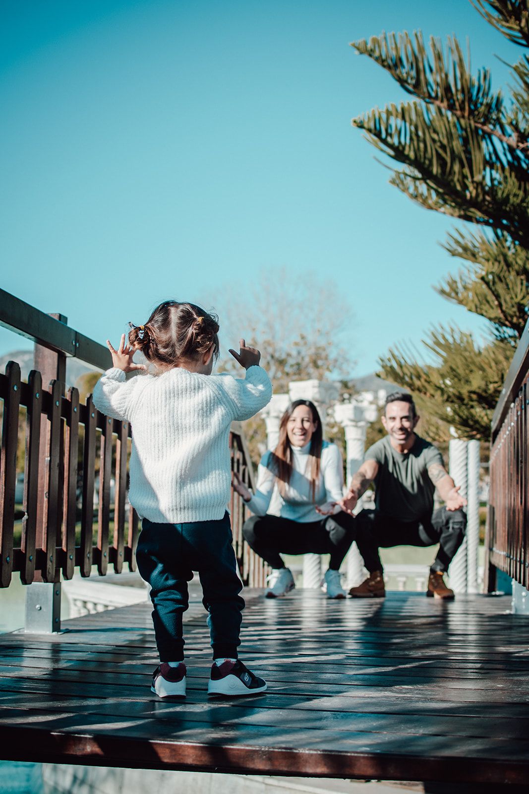 Un niño en una terraza de madera, con dos adultos agachados delante, al aire libre bajo un cielo azul