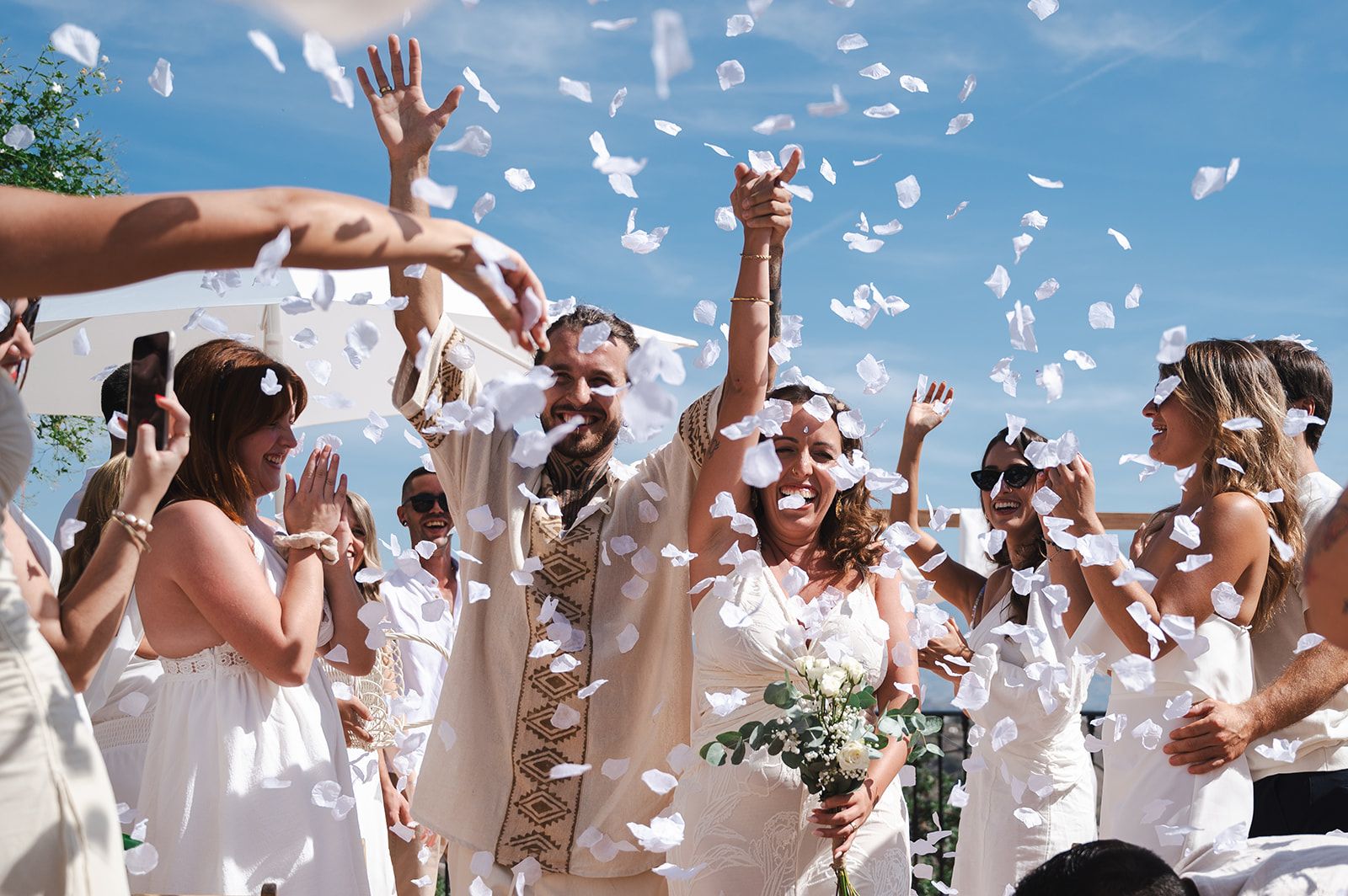 Los invitados de la boda, vestidos de blanco, celebran al aire libre mientras cae confeti bajo un cielo azul