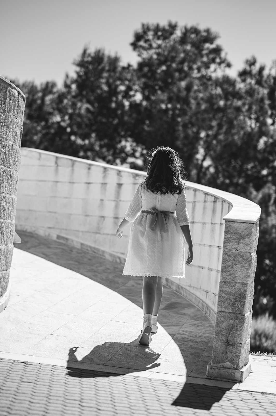 Mujer con un vestido blanco caminando junto a un muro de piedra curvo bajo un sol radiante