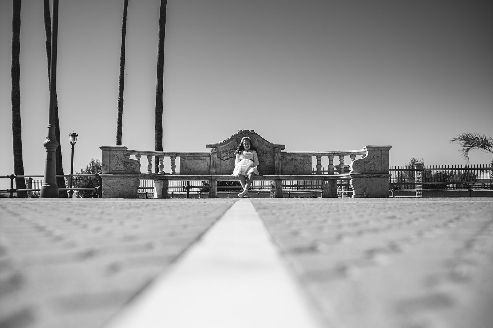 Vista en blanco y negro de un banco frente a la playa y palmeras, con un sendero centrado que lleva hasta el agua.