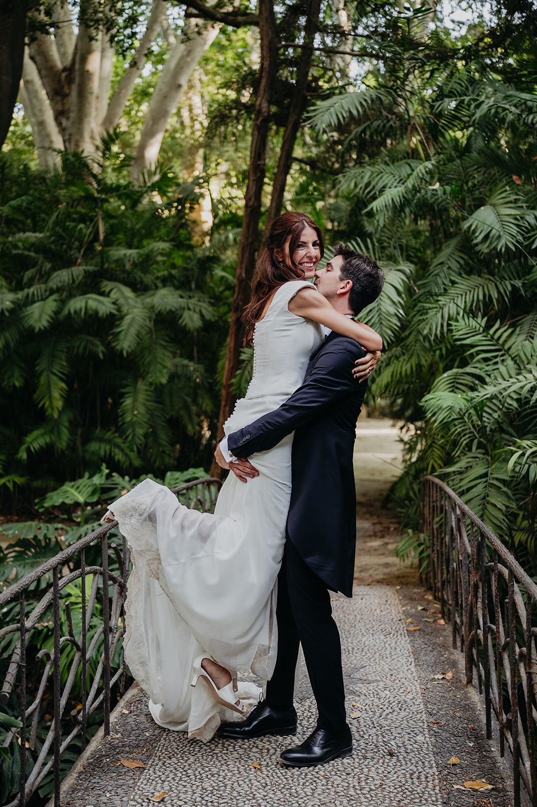 Una pareja vestida de novios se abraza en un sendero del jardín, rodeada de abundante vegetación