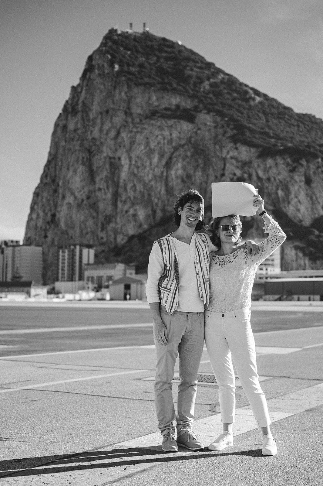 Dos personas de pie en una plaza, posando frente a una gran formación rocosa; una de ellas saluda con la mano.