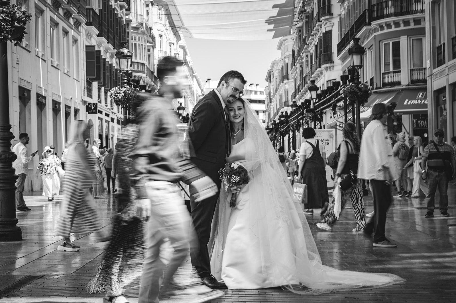 La novia y el novio se besan en una concurrida calle de la ciudad, mientras los peatones aparecen desenfocados por el movimiento a su alrededor.