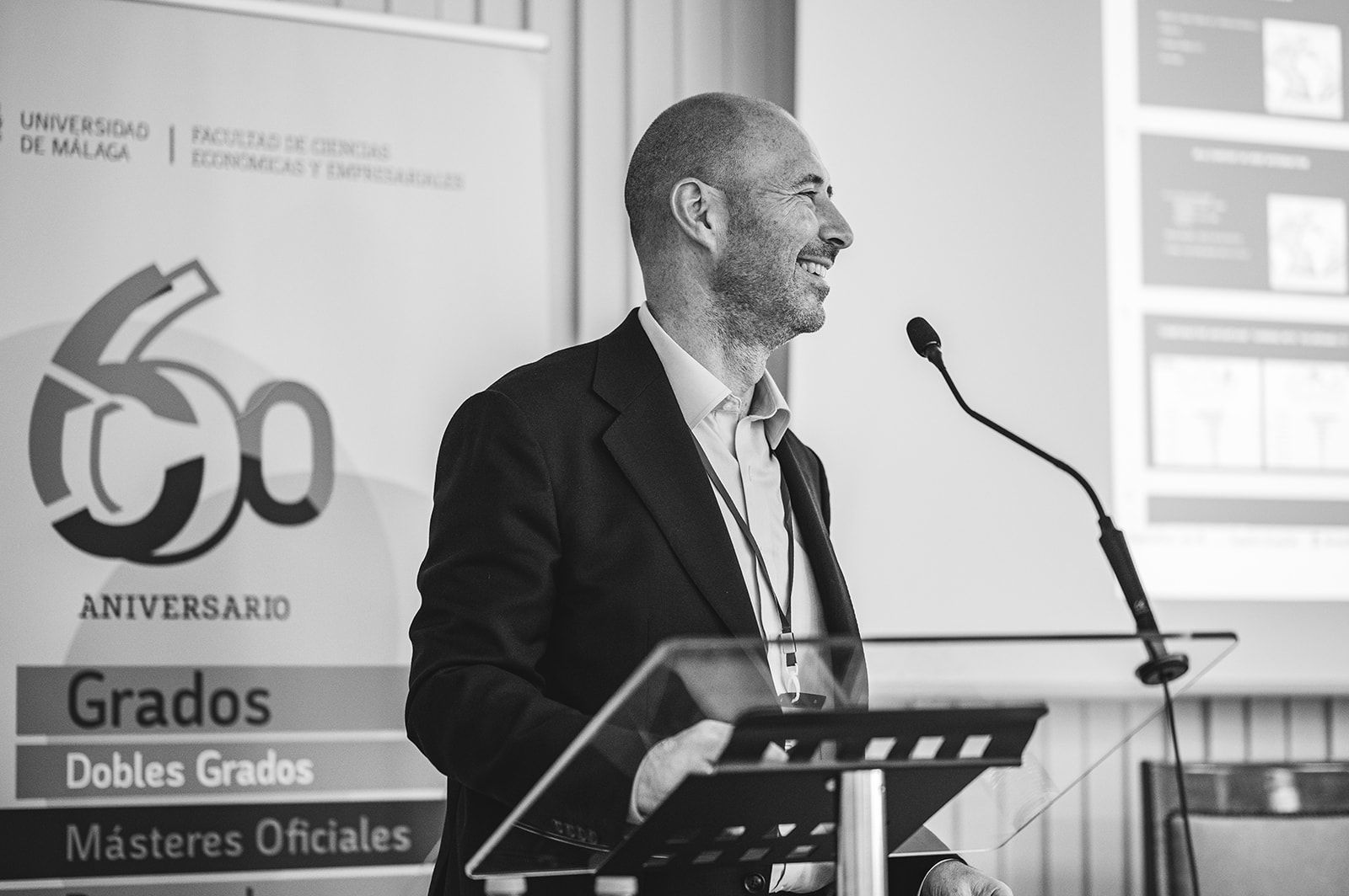 Un hombre habla desde un atril durante un acto académico por el 60.º aniversario, con una pancarta de titulaciones al fondo