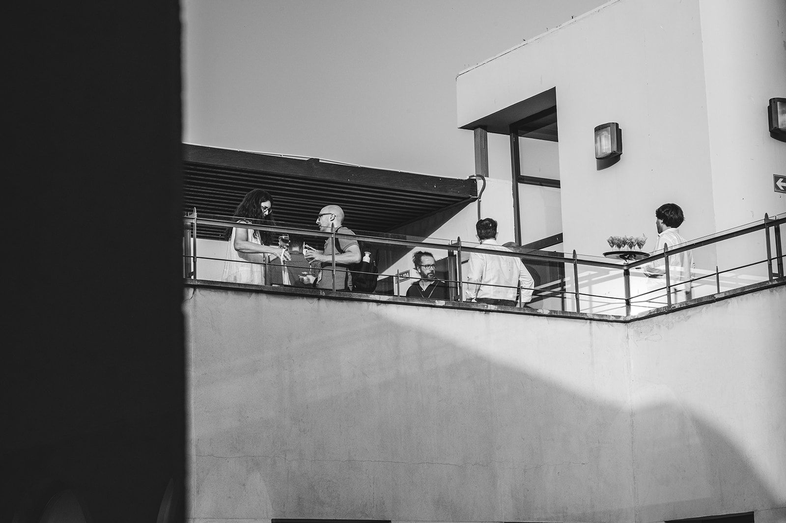 Vista en blanco y negro de personas en un balcón en la azotea, junto a un edificio blanco y una barandilla