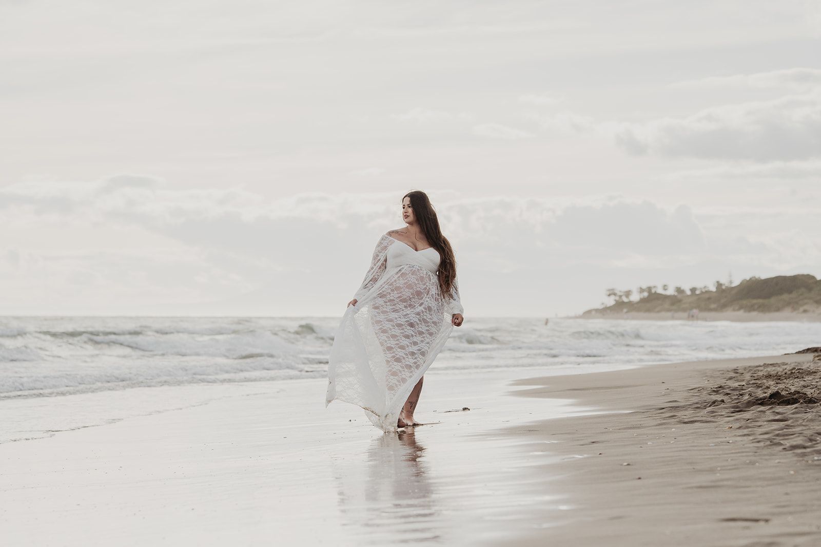 Una persona con un vestido blanco camina por una playa tranquila, cerca de la orilla.
