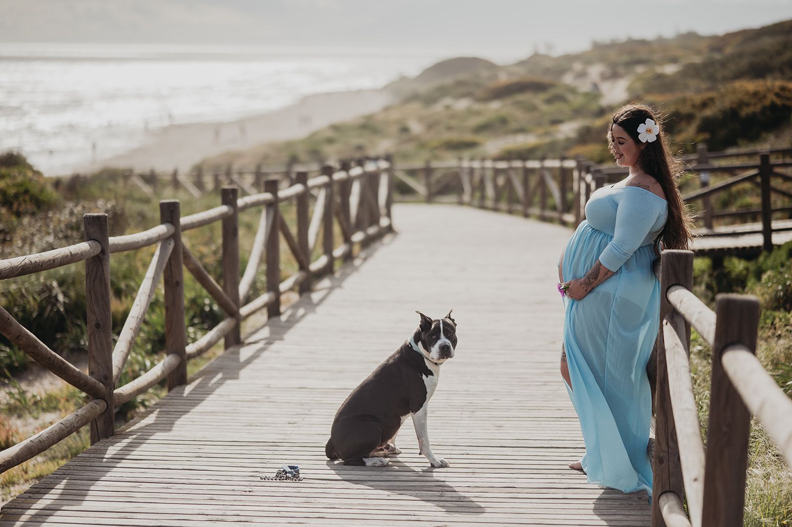 Mujer embarazada con un vestido azul de pie en una pasarela de madera, con un perro sentado a su lado cerca de la playa