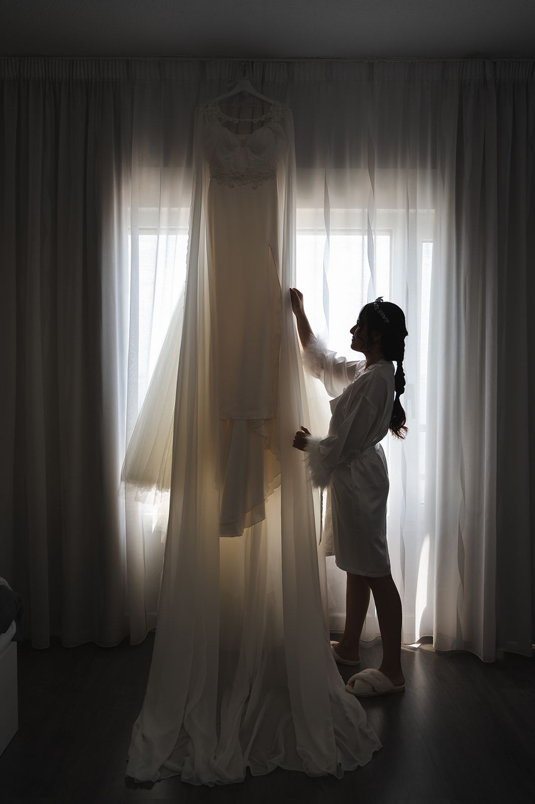 La novia ajusta un vestido de novia que cuelga junto a una ventana luminosa