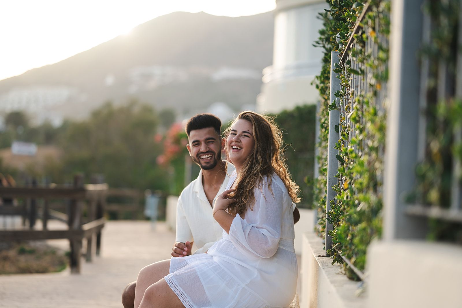 Pareja sonriente vestida de blanco, sentada al aire libre junto a una pared iluminada por el sol y rodeada de vegetación