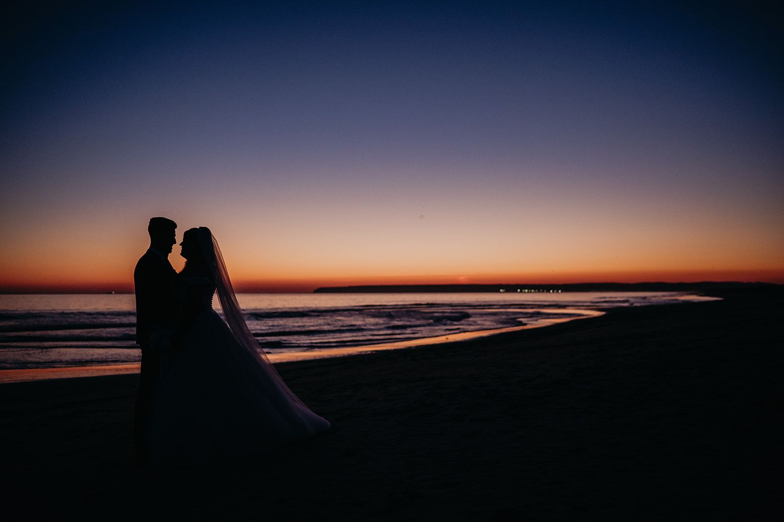 Una pareja en silueta de pie en una playa al atardecer, junto a las olas tranquilas y un cielo que se oscurece